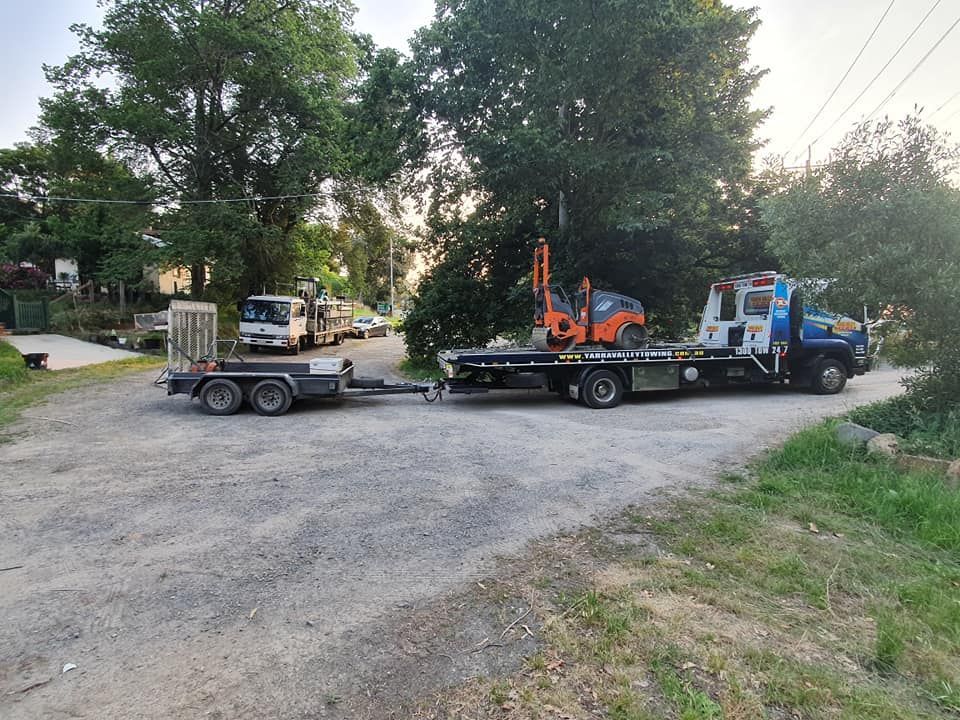 A tow truck with a small excavator on its bed, pulling a trailer on a gravel road.