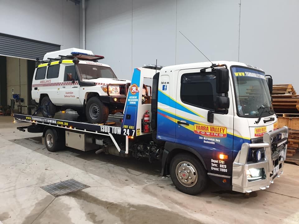 Tow truck with a white and yellow SUV on its flatbed, inside a warehouse.