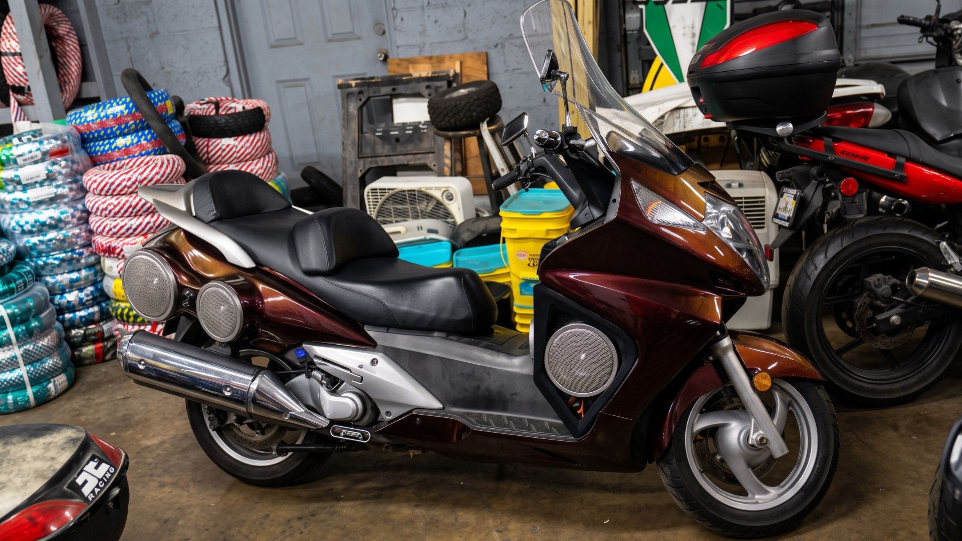 Brown touring scooter parked indoors among stacked goods and other motorcycles.
