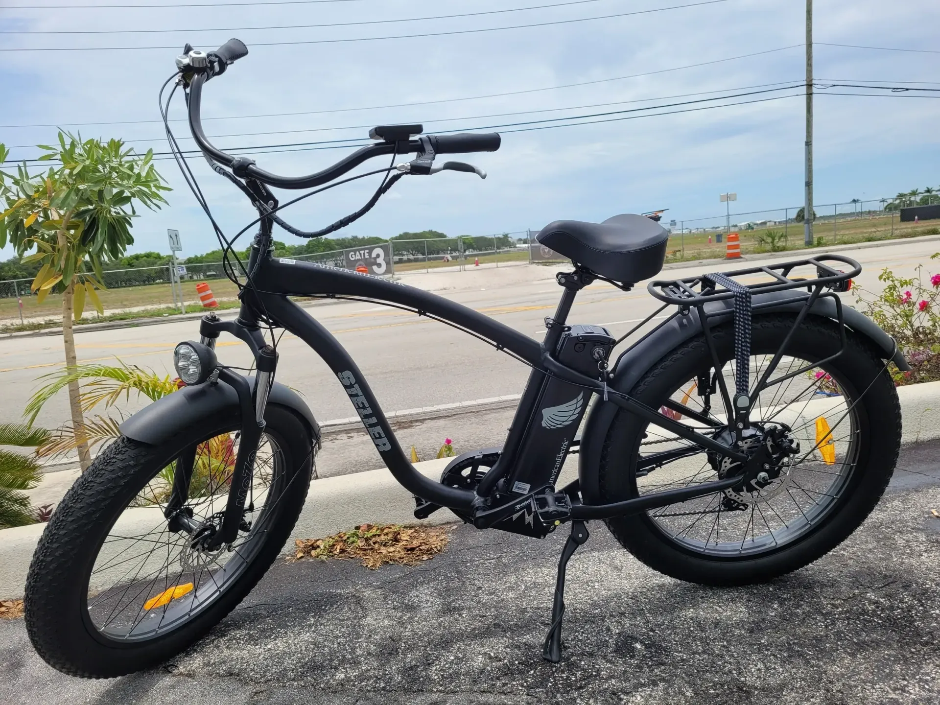 Black electric bicycle parked by the roadside under a cloudy sky, with a rear rack and wide tires