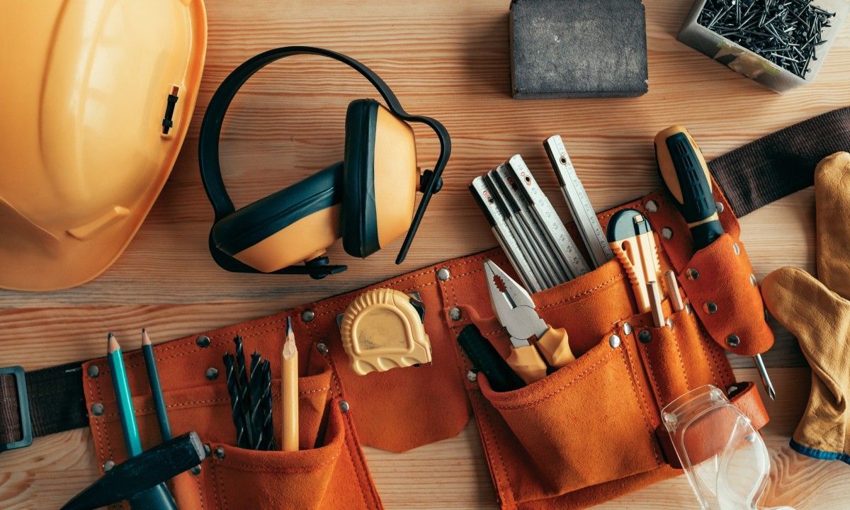 A close-up of a handyman’s toolbelt filled with various tools sits on a table next to a hard hat, a sanding pad, and nails.