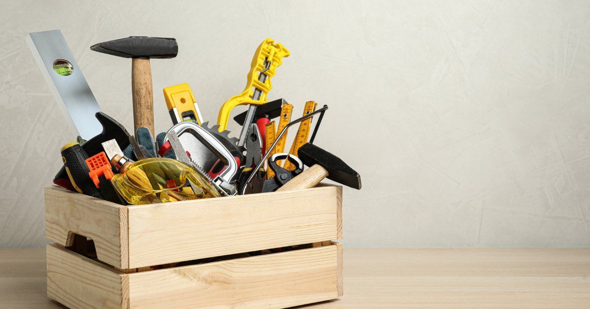 A wooden crate sits on a plain work surface against a white backdrop. The crate is full of handyman tools.
