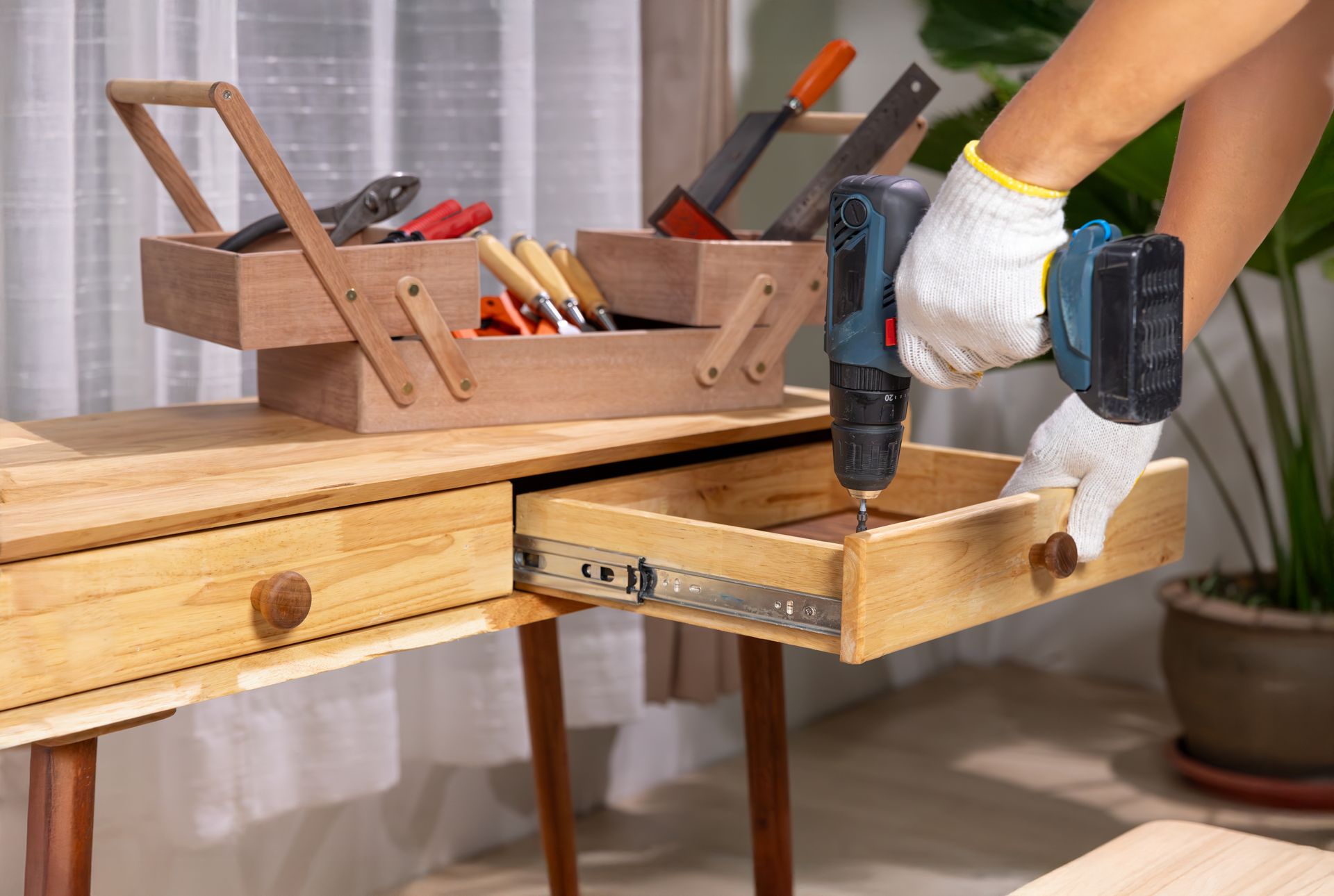 Person with glove using a drill on a drawer while wooden toolbox sits on a table.