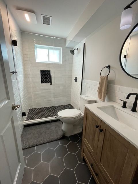A bright bathroom features a walk-in shower with dark hexagonal floor tiles, a white toilet, and a wooden vanity.
