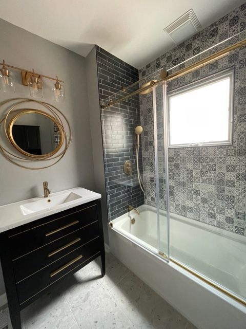 A modern bathroom with a black vanity, gold fixtures, a circular mirror, and a tub with patterned and subway tile walls.