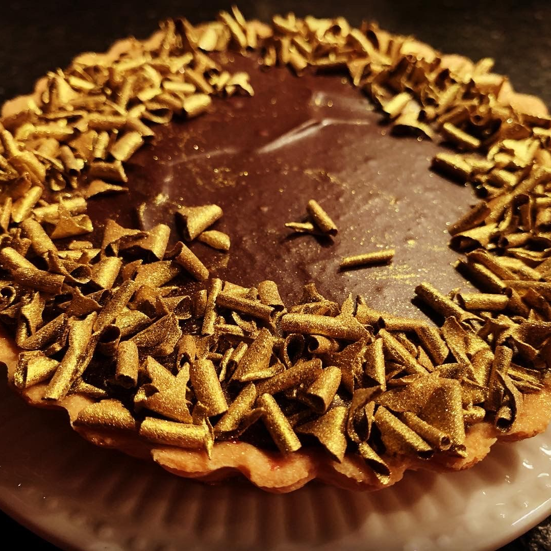 A chocolate tart in a fluted crust, garnished with a ring of metallic gold shavings around the dark filling.