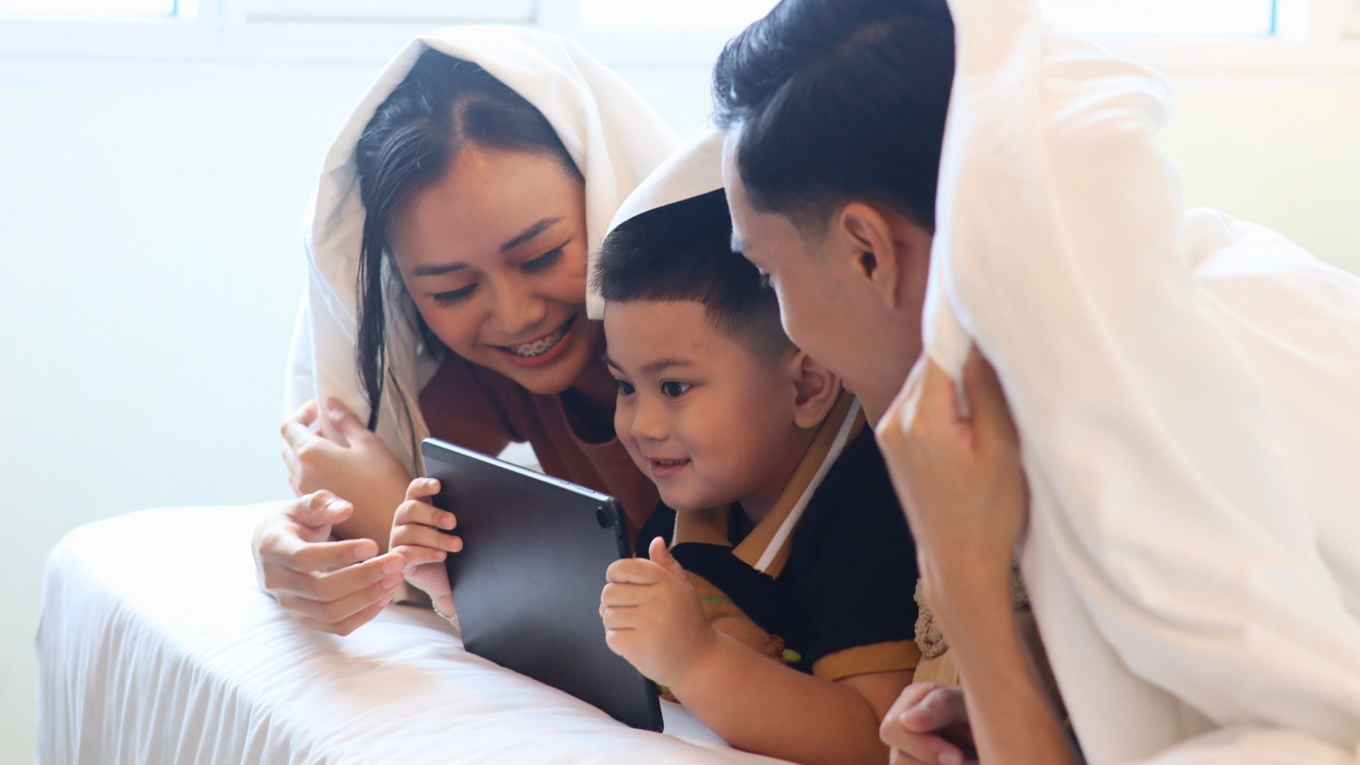 Family under a white blanket smiling and looking at a tablet on a bed