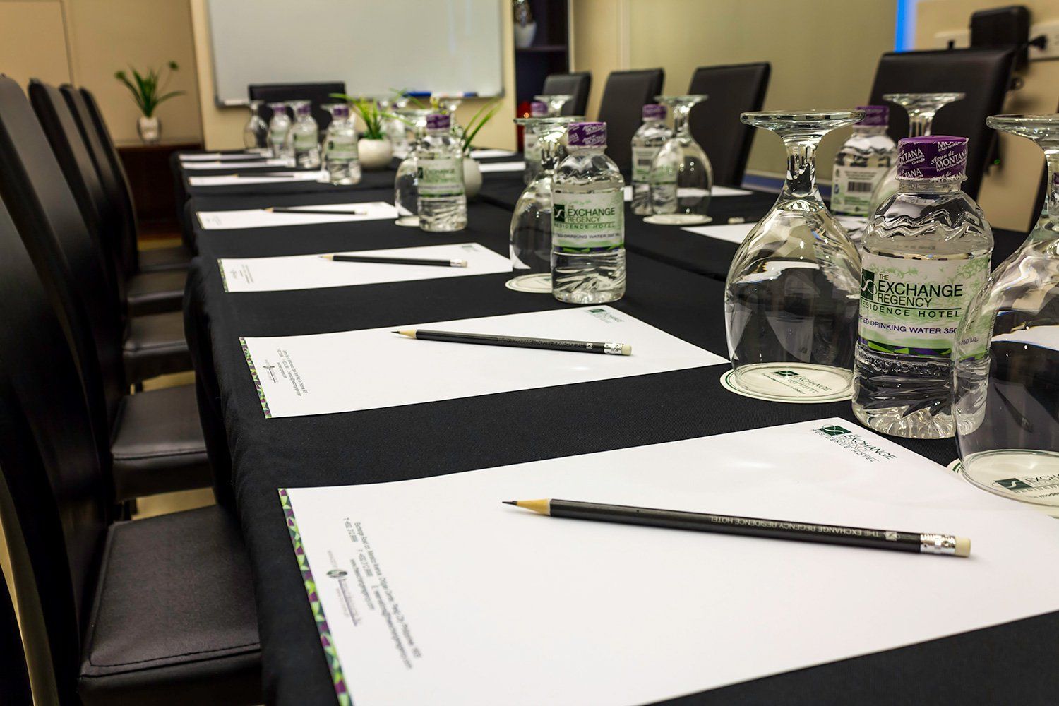 Conference table set with water bottles, glasses, notepads, and pencils. Black tablecloth, chairs in a room.