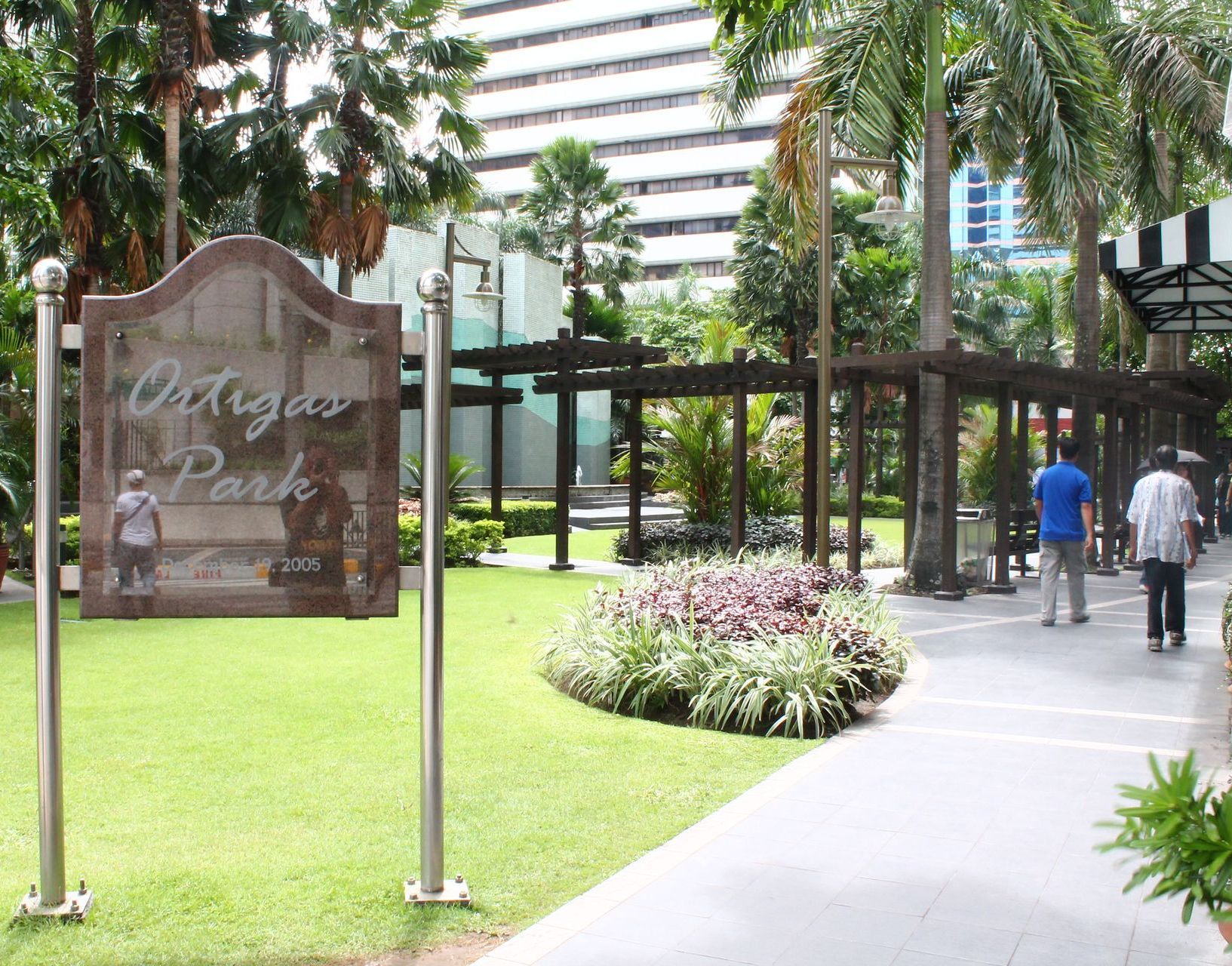 Park entrance sign beside a walkway and lawn, with people walking near palm trees and a high-rise building.