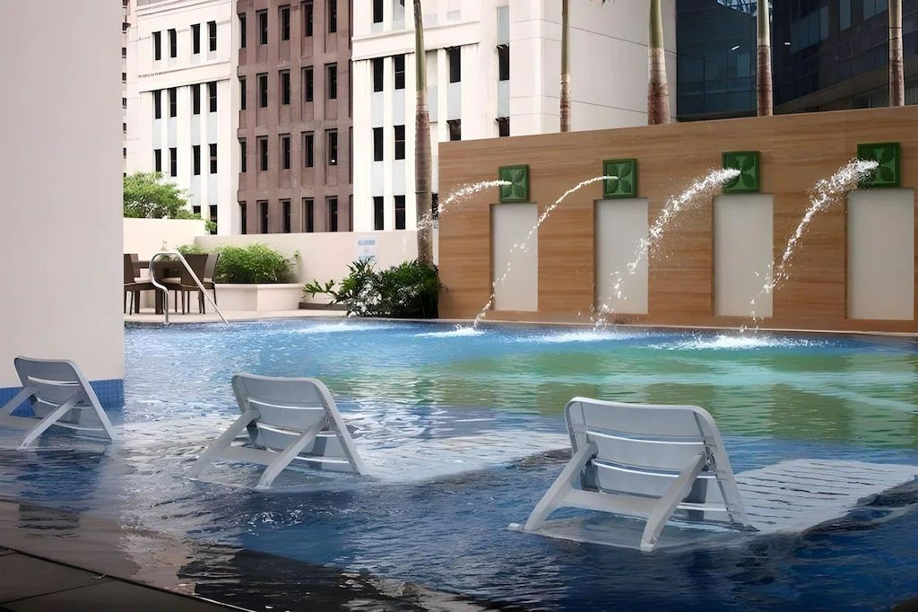 Pool with chairs in the water, fountains, and buildings in the background.