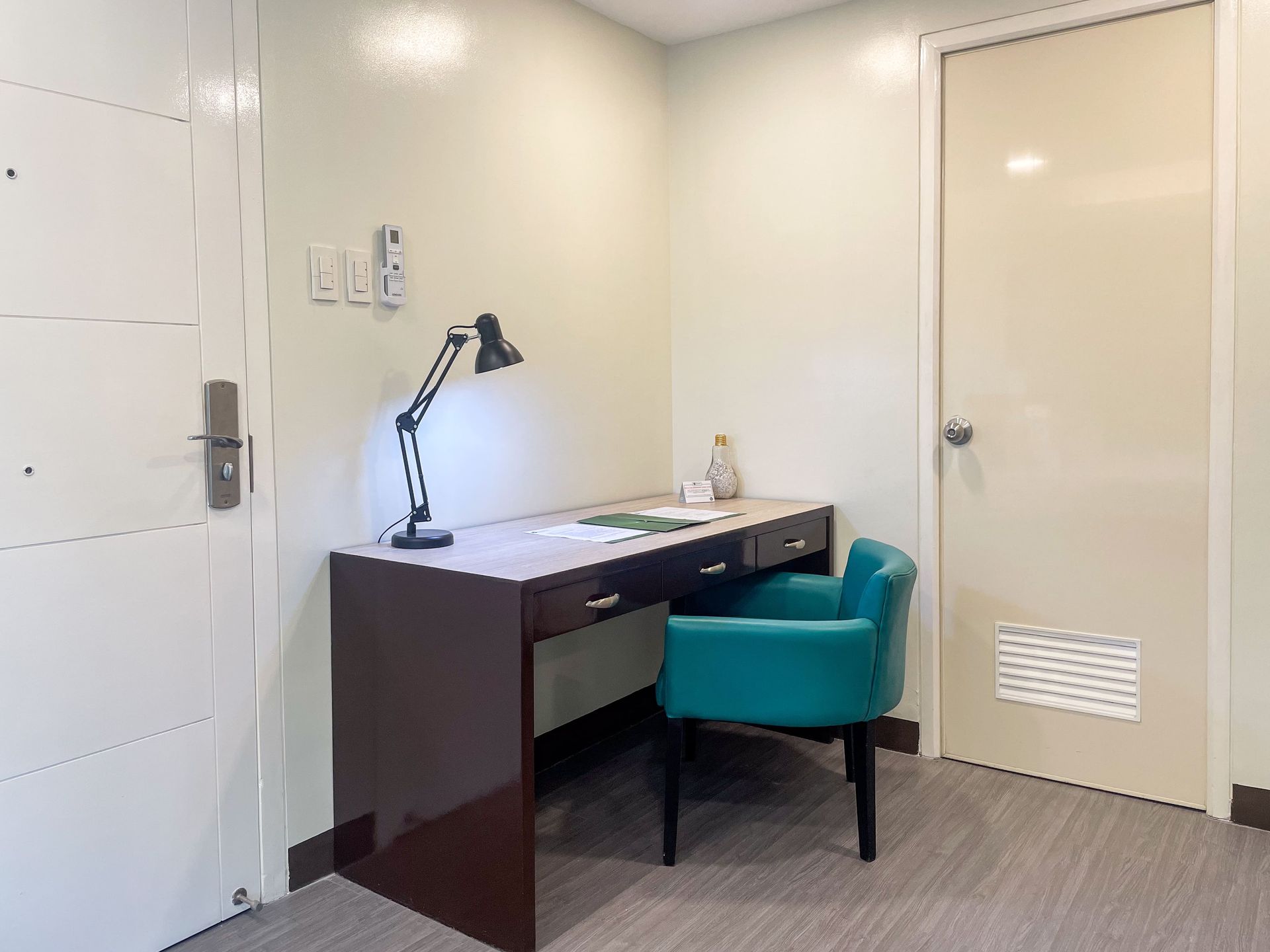 A hotel room with a desk , chair , lamp and door.