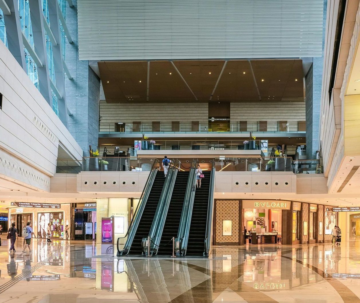 Mall interior with escalators and shops. Light flooring, high ceiling, and people shopping.