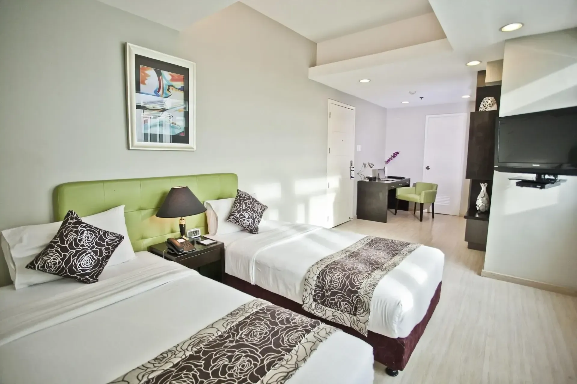 Twin beds in a modern hotel room; green headboard, desk, TV, artwork, and light-colored walls.
