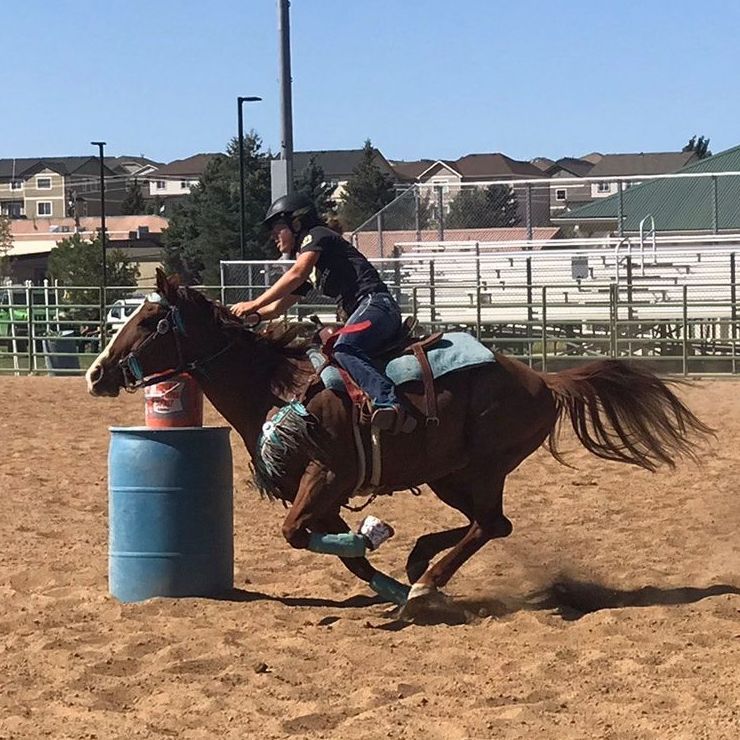 Woman riding a brown horse in a barrel racing competition, turning around a blue barrel in a sandy arena.