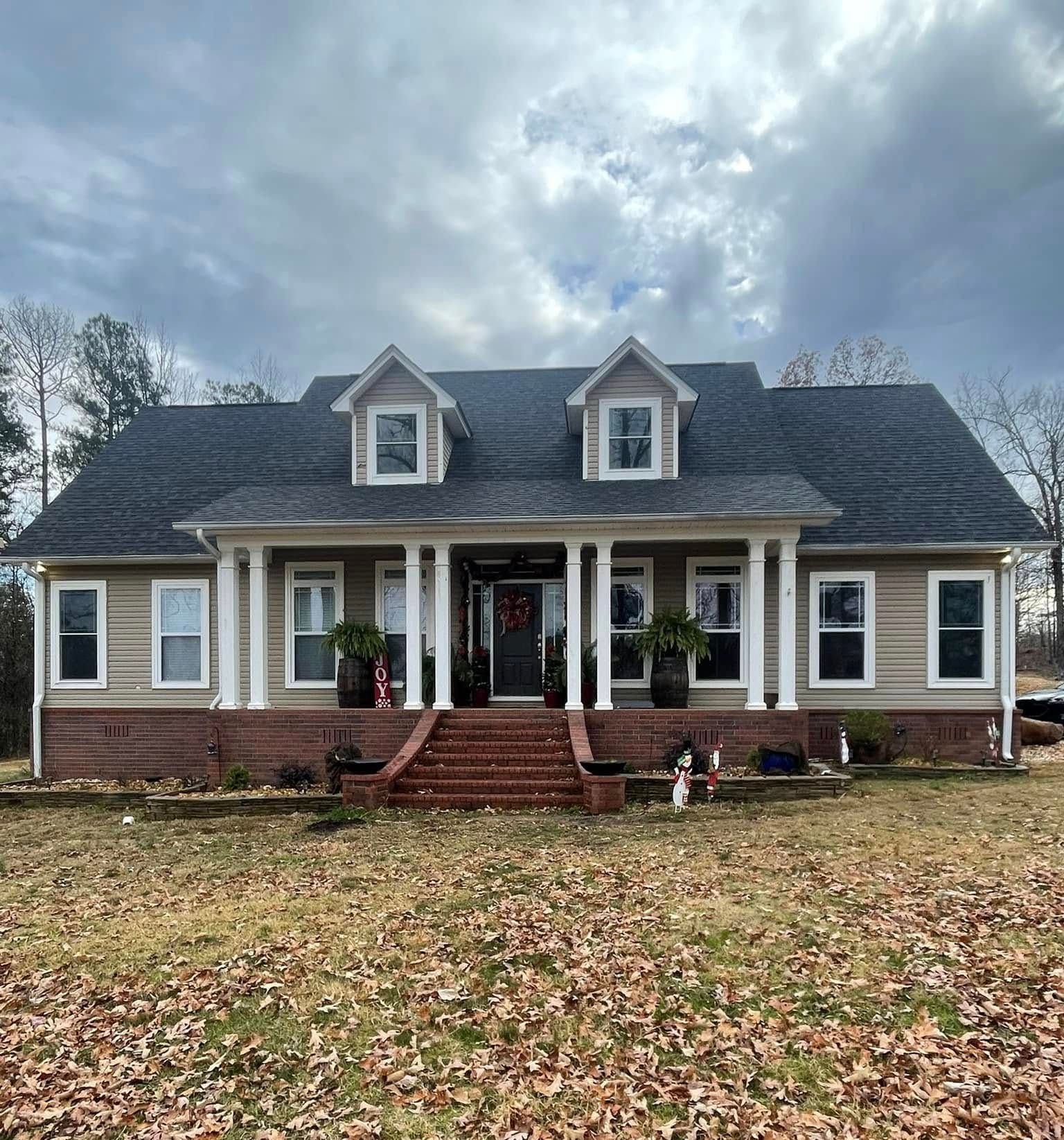 Tan house with a porch, brick steps, and two dormers under a cloudy sky.