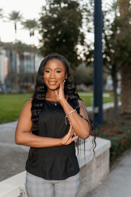 Woman in a black top and patterned pants stands smiling outdoors by a low wall.