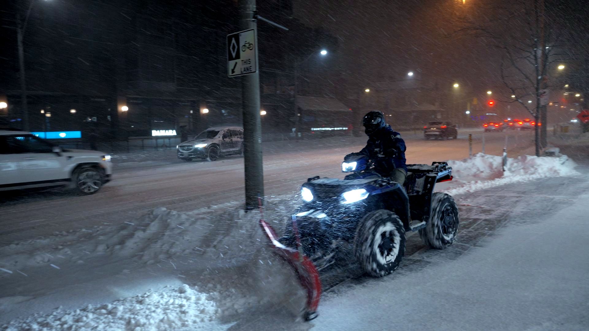 An ATV with a snowplow clearing a sidewalk on a snowy street at night.