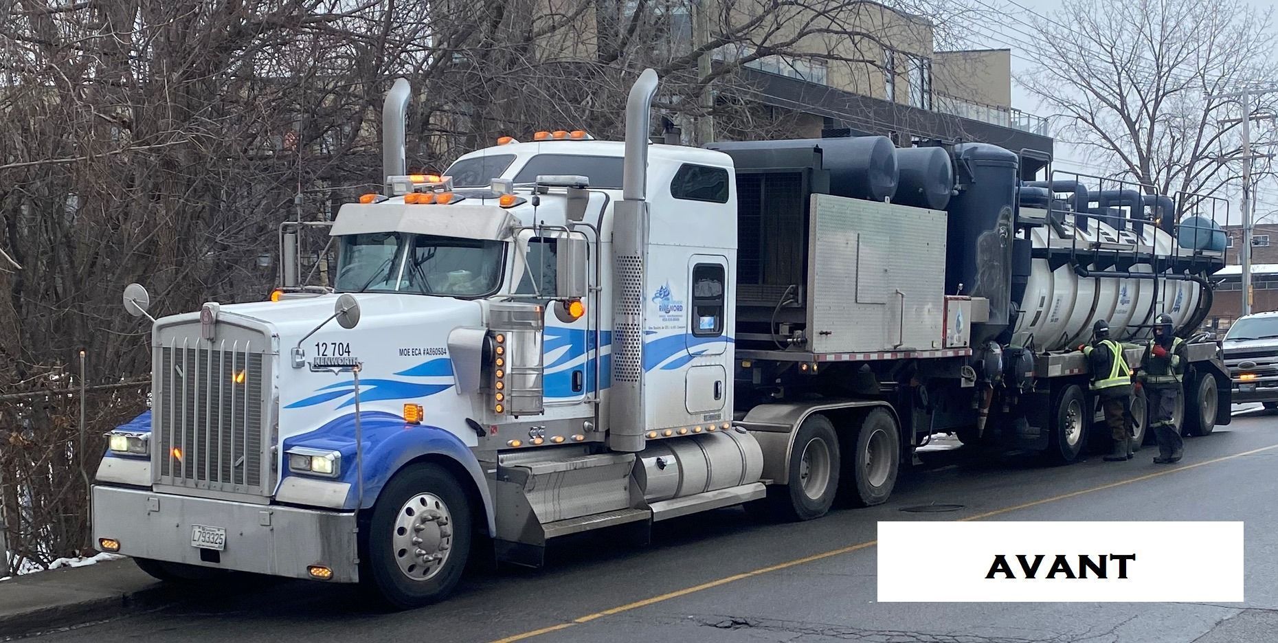 Un gros camion blanc avec une remorque-citerne est garé dans la rue. Deux personnes se tiennent à l'arrière.