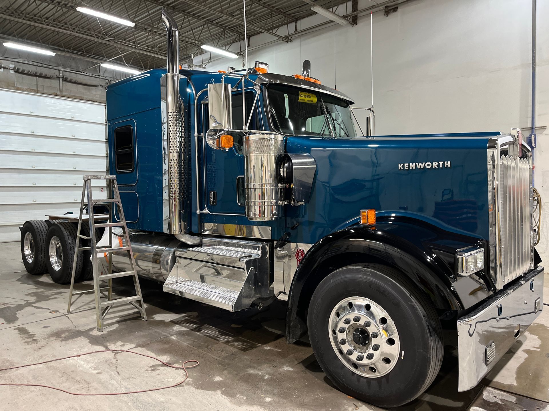 Un semi-remorque Kenworth bleu foncé est garé dans un garage intérieur, avec un escabeau métallique à côté.