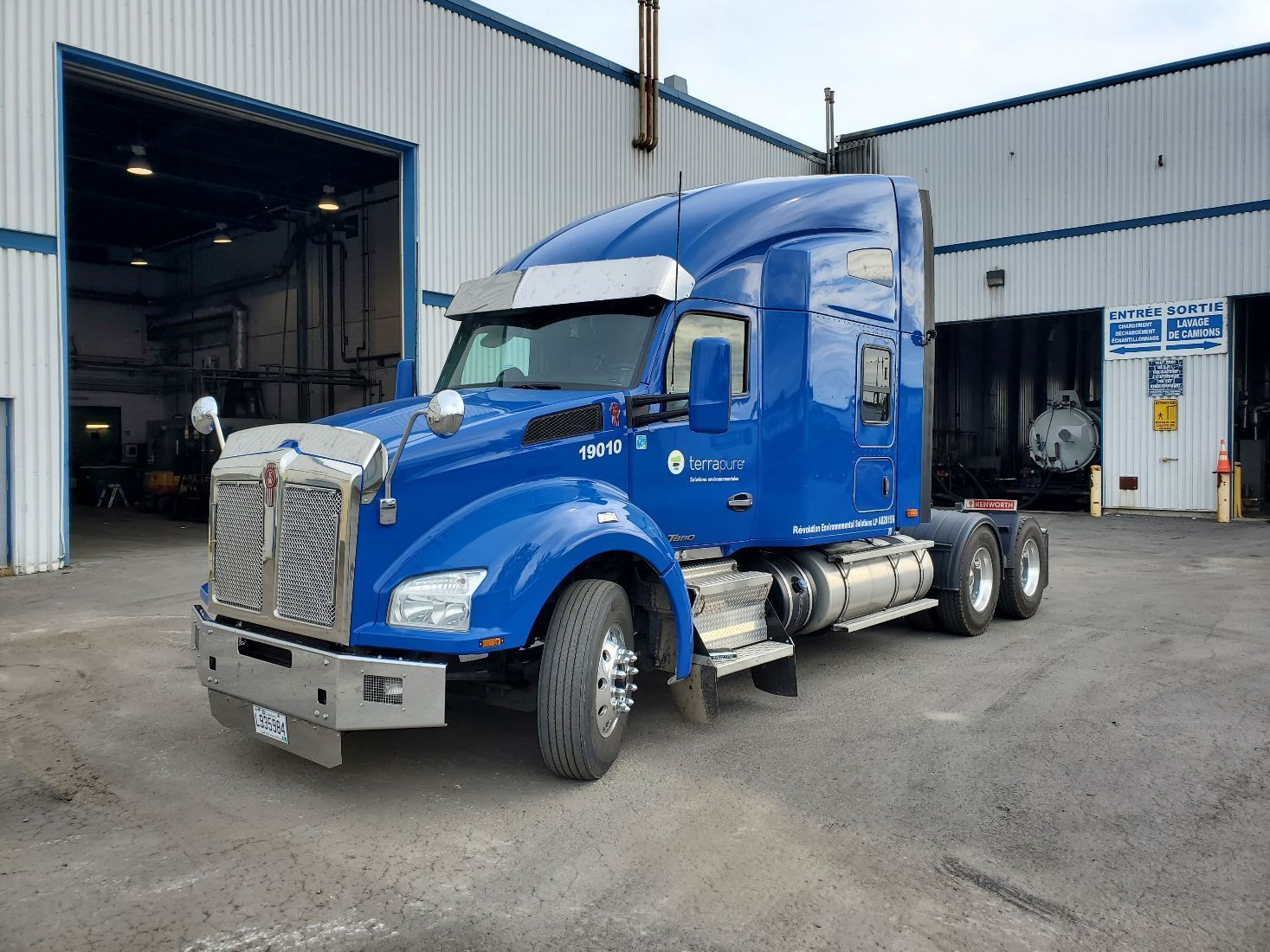 Semi-camion bleu Kenworth garé à l'extérieur d'un entrepôt avec la porte ouverte, à la lumière du jour.