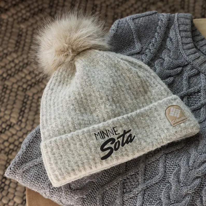 A white beanie with a pom pom is sitting on top of a gray sweater.