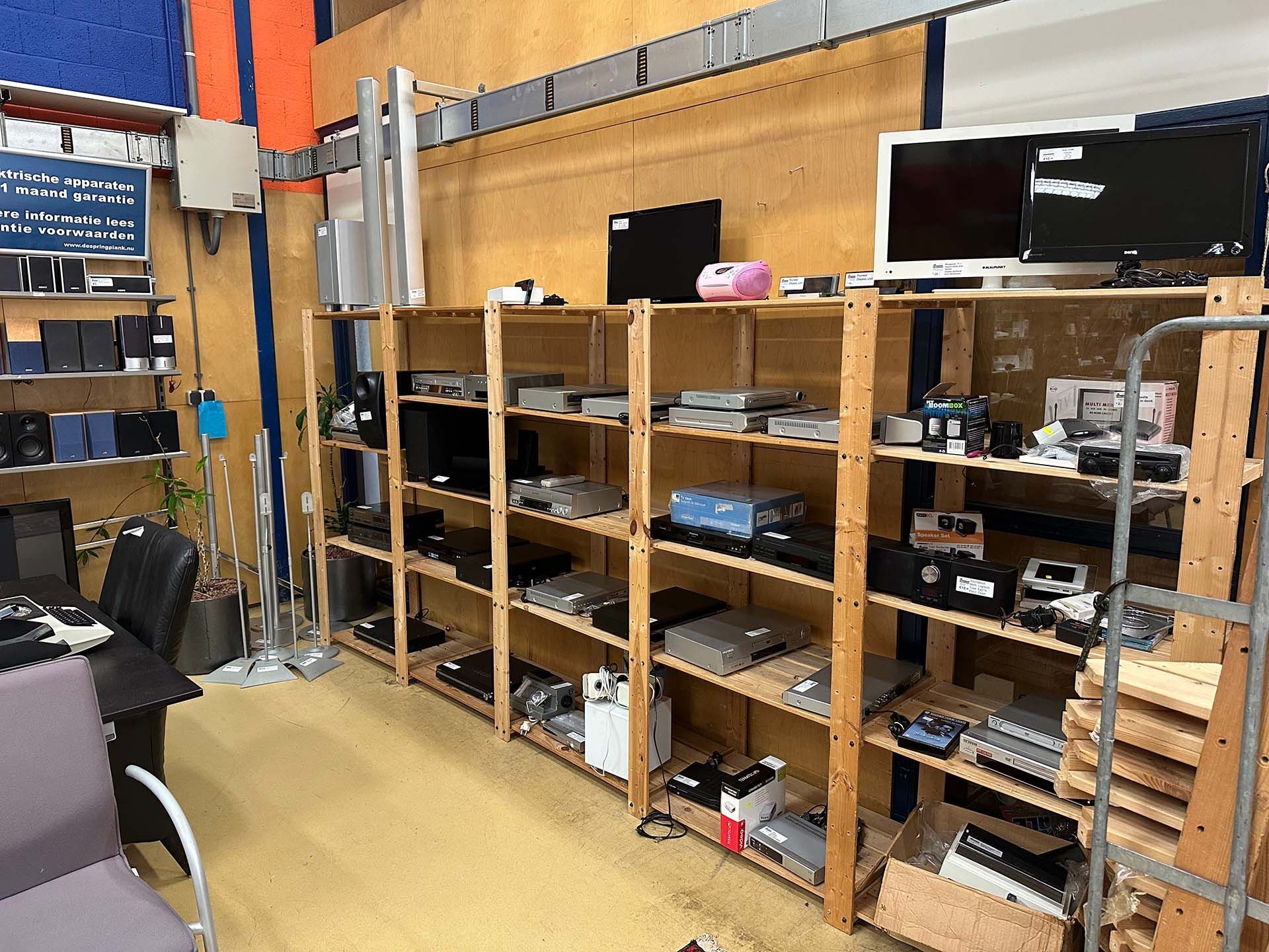 Schappen vol elektronische apparatuur en onderdelen in een werkplaats, met monitoren en gereedschap op houten rekken.