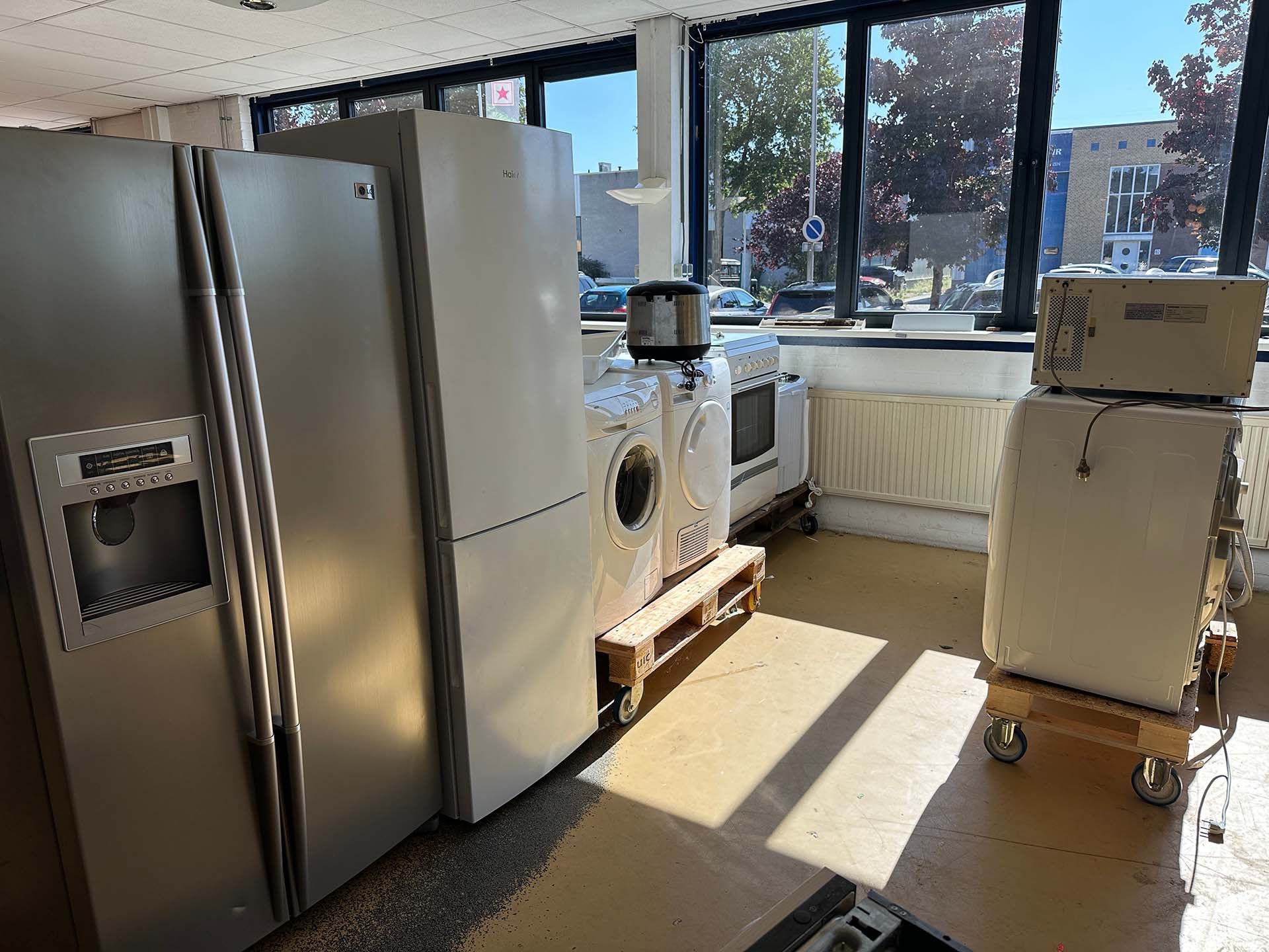 Keuken met roestvrijstalen apparaten, ramen waar het zonlicht doorheen schijnt en gestapelde dozen op een kar.