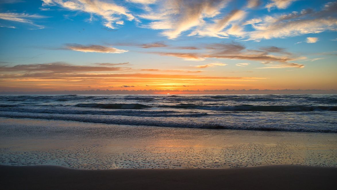 Sunset over the ocean, with waves crashing on the beach. Orange and blue sky, reflecting on the wet sand.