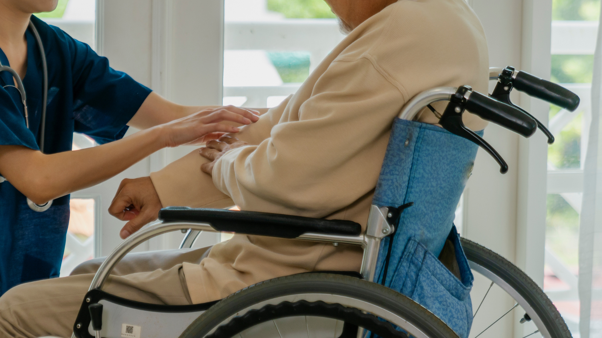 Nurse assisting a man in a wheelchair. The nurse touches the man's arm in a room with a window.