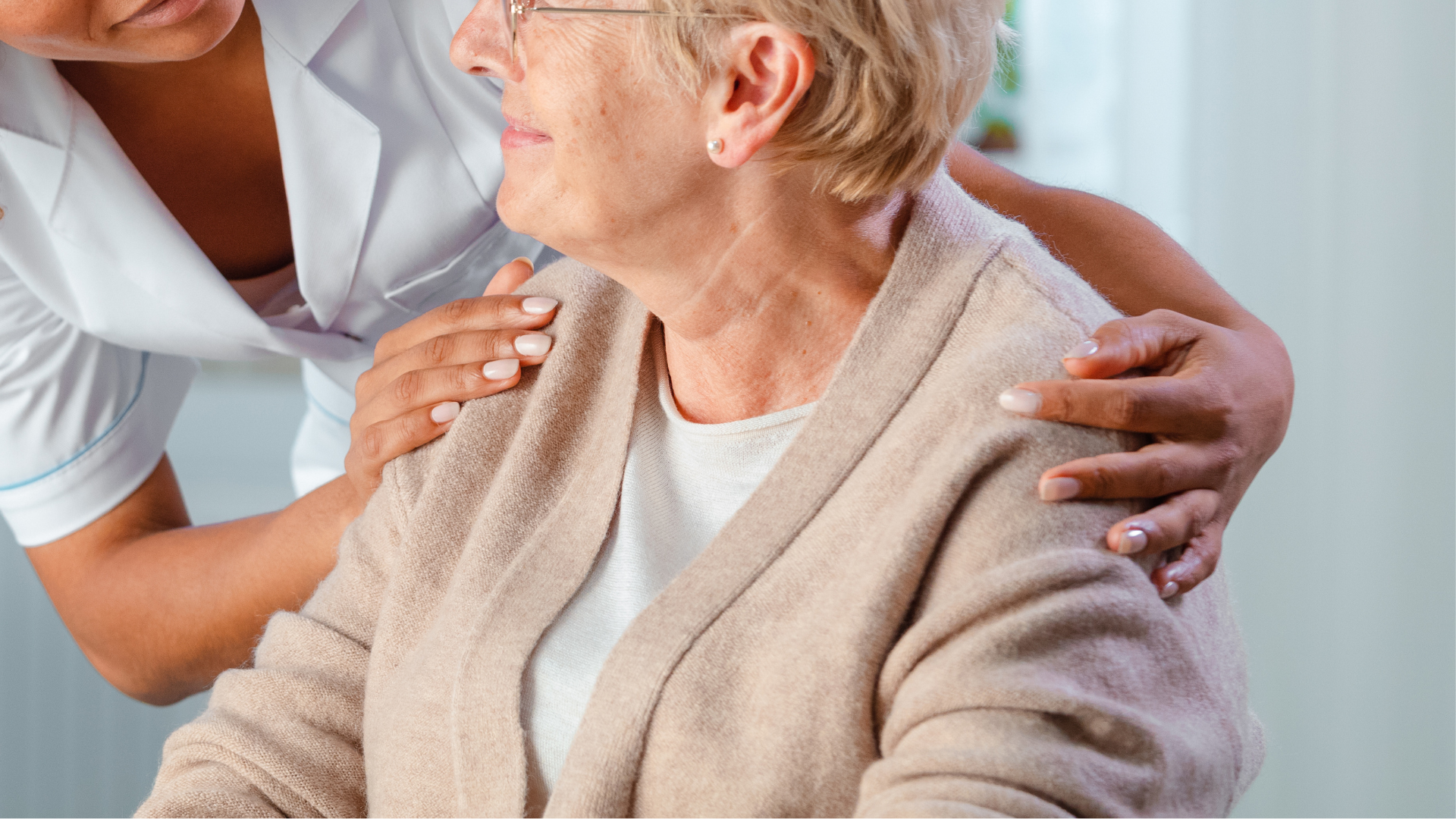 Nurse's hands on the shoulders of an elderly woman. Both smiling, indoors.