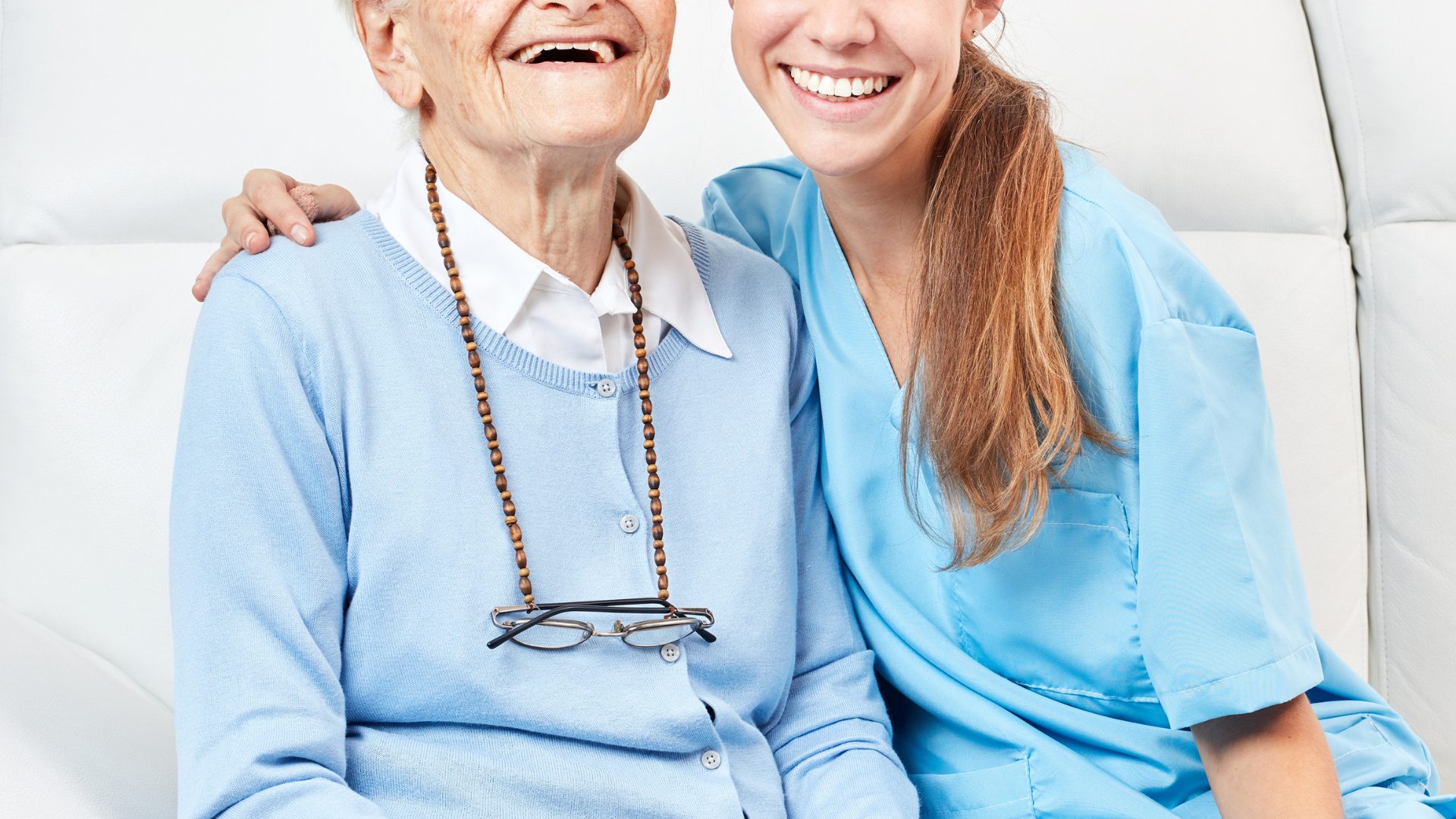 Elderly woman with caregiver, both smiling on a white couch. Caregiver has an arm around the woman.