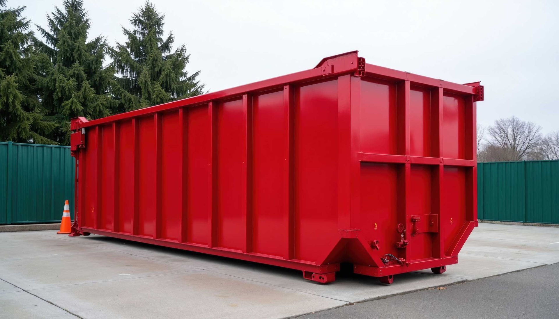 Large red industrial dumpster on a concrete lot with green fencing and trees in the background Large red industrial dumpster on a concrete lot with green fencing and trees in the background