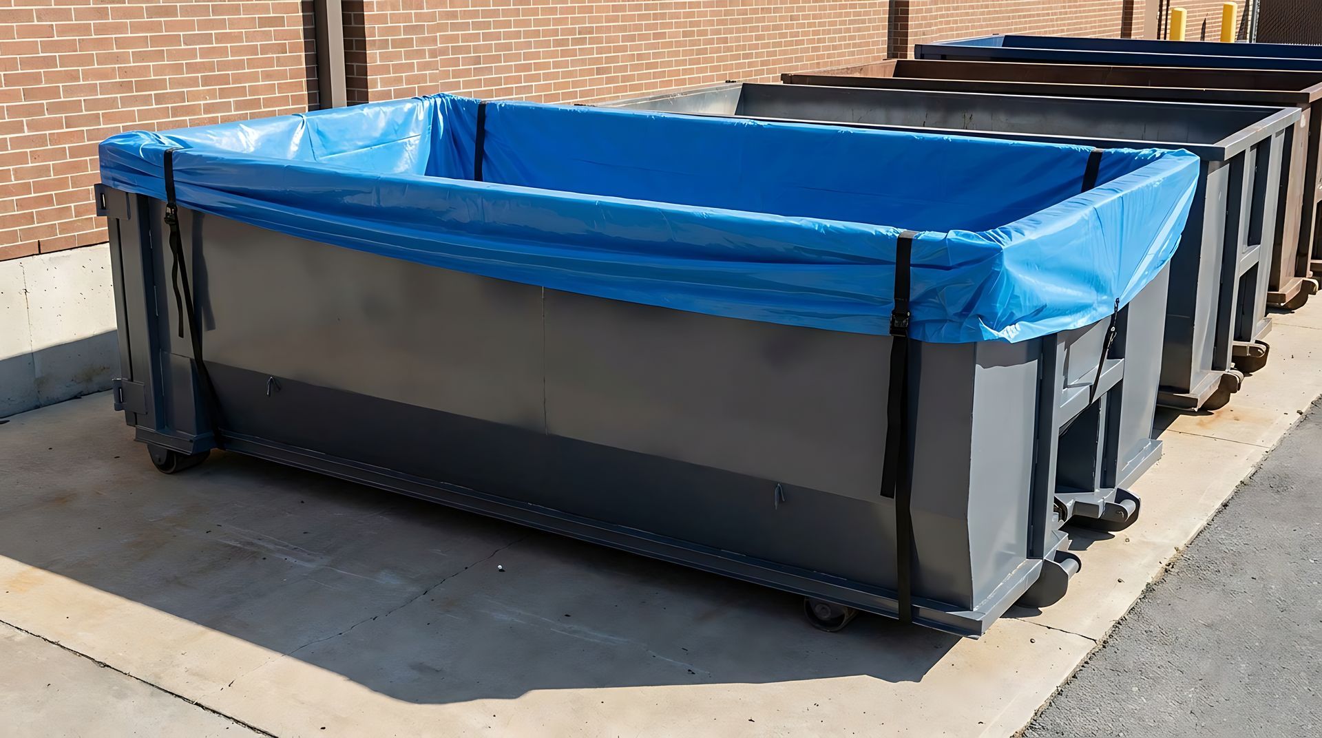 A large metal roll-off dumpster fitted with a bright blue tarp liner rest on a paved lot outdoors. A large metal roll-off dumpster fitted with a bright blue tarp liner rest on a paved lot outdoors.
