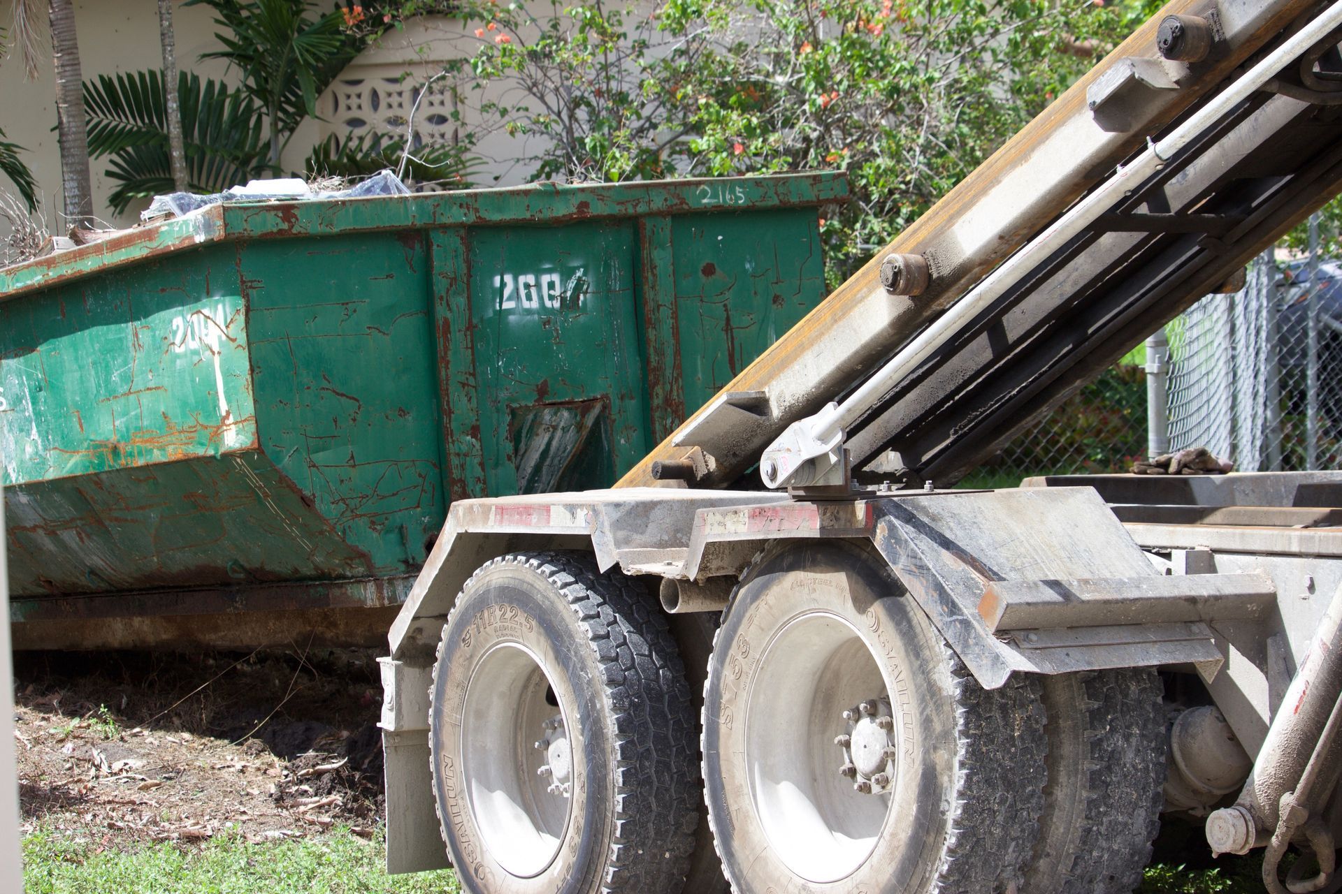 Green construction dumpster beside a truck loading debris in a residential yard.