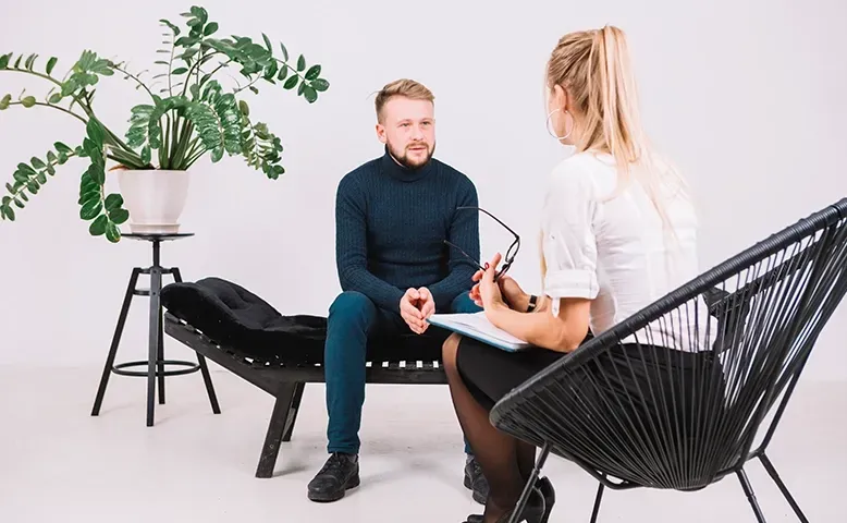 Two people talking in a bright office lounge, seated in black modern chairs near a potted plant.