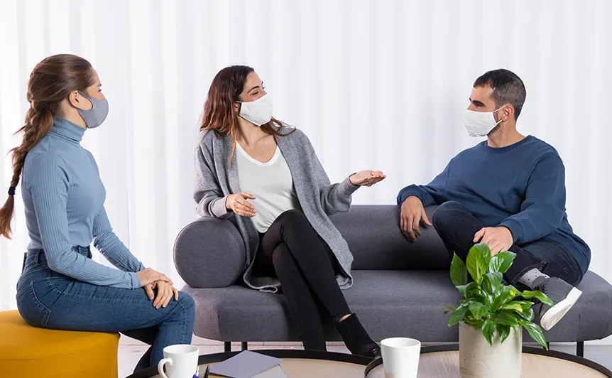 Three masked people sit in a circle talking during a counseling session with coffee mugs nearby