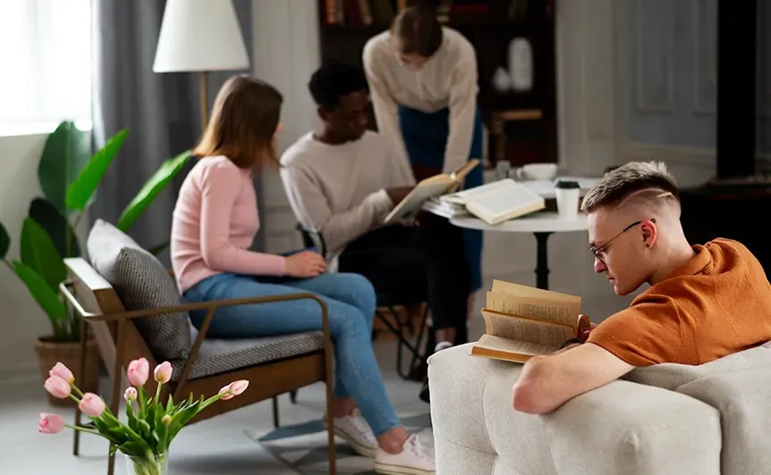 People chatting in a modern living room, with one person reading from a book and others seated nearby.
