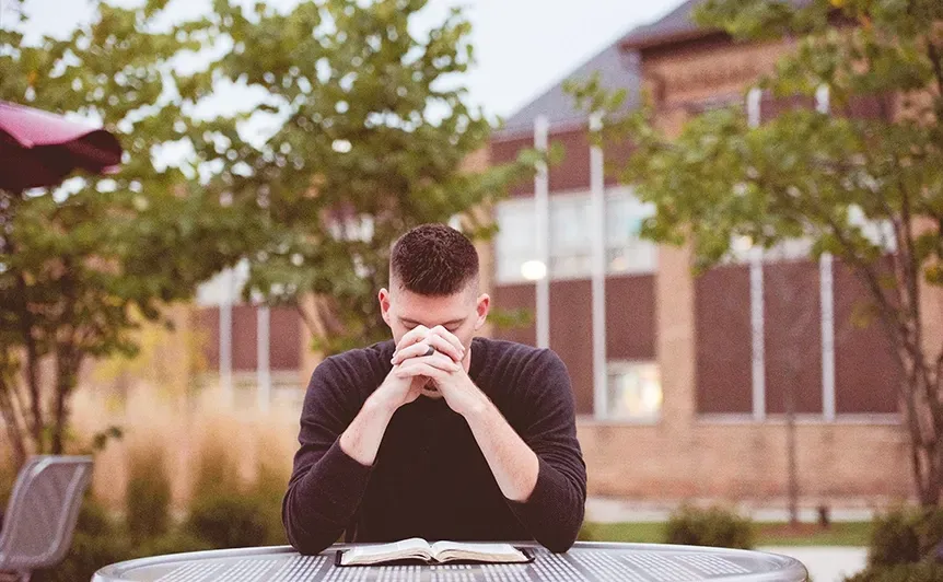 Man sitting at an outdoor table with head bowed and hands clasped, in front of a building and trees
