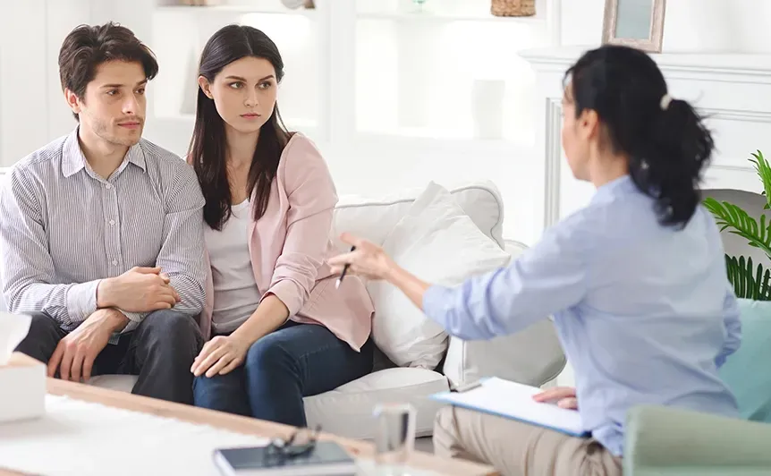 Two people listening to a counselor in a bright living room, seated on a sofa during a therapy session