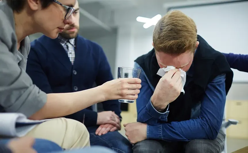 Three people in an office: one wipes tears while others comfort him, one offering a glass of water.