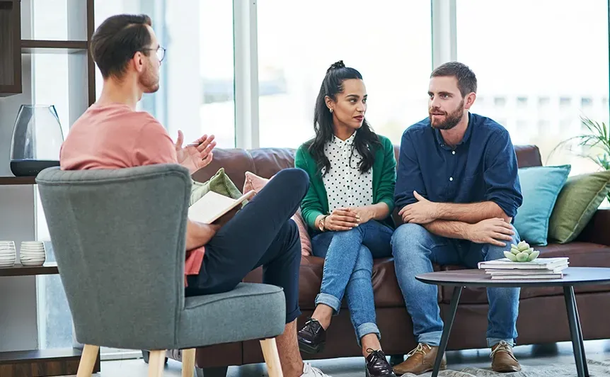Three people in a living room having a conversation, sitting on chairs and a sofa near a coffee table.