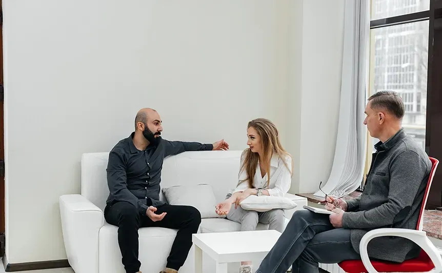 Three people in a bright office meeting, seated on white couches and discussing papers.