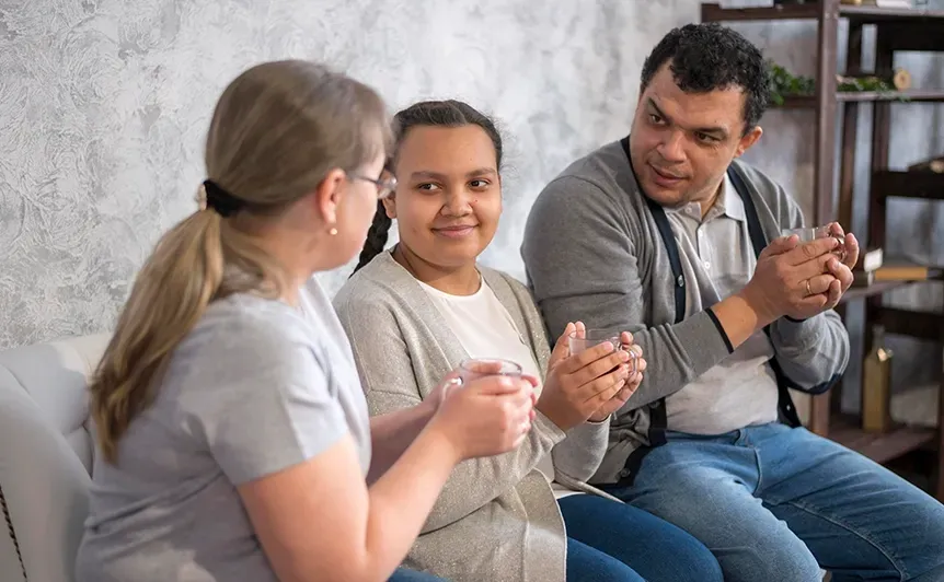 Three people sit on a couch, smiling and talking in a bright living room.