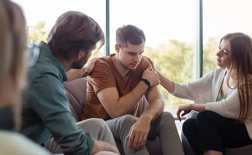 Support group meeting with three people talking in a bright room, one consoling another on the shoulder.