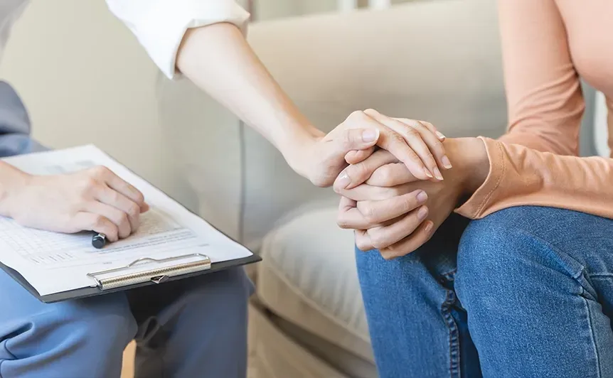 Two people holding hands during a counseling session, with a therapist taking notes nearby.