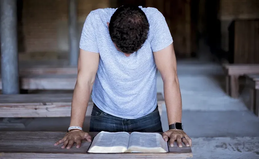 Person bowing over an open book in a dim room, hands on the floor in a prayer posture.