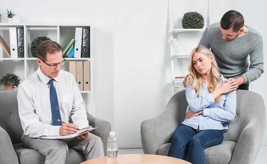 Counselor with notepad talks to a seated woman while a man stands beside her in a bright office