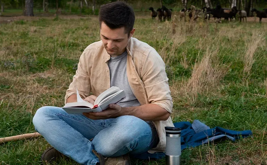Man sitting cross-legged on grass, reading a book beside a water bottle and backpack outdoors.