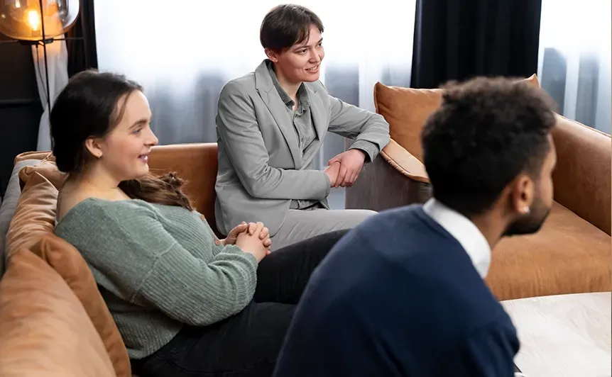 Three people sit on a couch in a living room, listening attentively during a discussion.