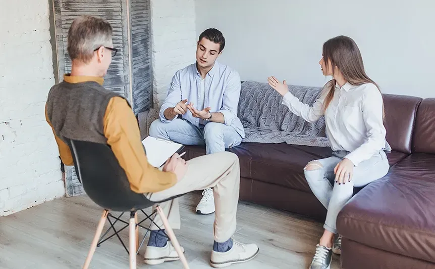 Three people in a living room discussing, seated on a sofa and chairs